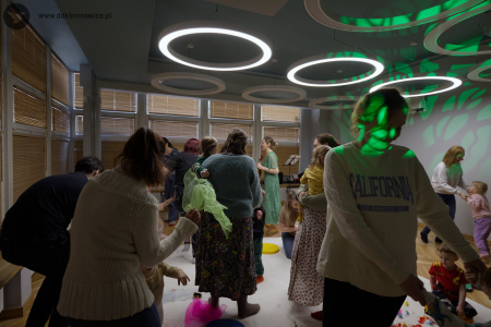 Rodzice tańczą ze swoimi dziećmi. Na suficie okrągłe lampy. Po prawej stronie projekcja drzewa.