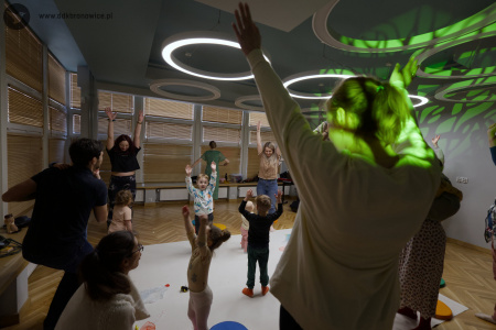 Dzieci i dorośli tańczą na białym dywanie. Uczestnicy podnoszą ręce do góry. Uczestnicy są uśmiechnięci i dobrze się bawią.