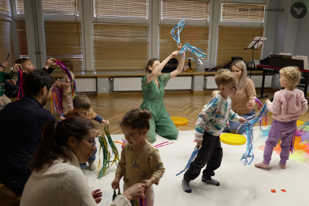 Dzieci bawią się na białym dywanie kolorowymi wstążkami. Prowadząca warsztaty siedzi na białym dywanie. Trzyma w rękach biało-niebieskie wstążki.