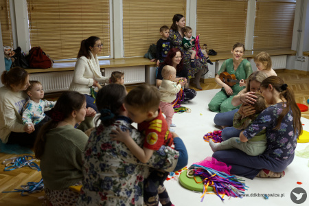 Uczestnicy siedzą na dywanie. Sylwia Namroży gra na ukulele. Dzieci odpoczywają po zabawie na nogach rodziców.