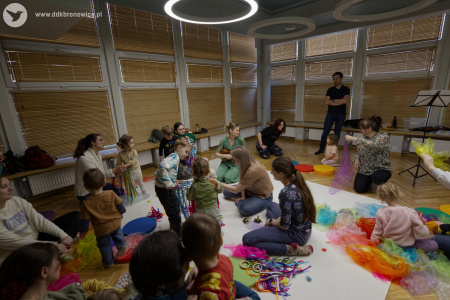 Dzieci bawią się szarfami. Po prawej stronie dziewczynka w różowej bluzie siedzi na kolorowych chustach. Prowadząca siedzi na białym dywanie i gra na ukulele.