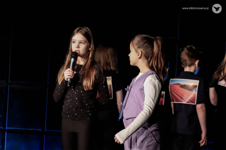 Dziewczynka ubrana na czarno mówi do mikrofonu. Trzyma go w prawej ręce. Obok niej stoi dziewczynka w fioletowym kostiumie.