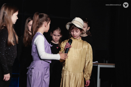 Scena teatralna. Dzieci stoją na scenie. Z przodu rozmawiają dwie dziewczynki. Dziewczynka po lewej ubrana w fioletową kamizelkę i spodnie. Dziewczynka po prawej w złotą sukienkę.