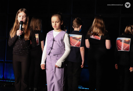 Na środku stoi dziewczynka ubrana w fioletowy kostium. Za nią stoją dzieci plecami do widowni. Na czarnych koszulkach widać krajobrazy pustynne. Po lewej stronie stoi dziewczynka ubrana na czarno. Mówi do mikrofonu.