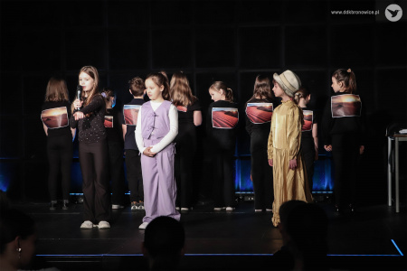 Z przodu stoją trzy dziewczynki. Od lewej dziewczynka ubrana na czarno, dziewczynka w fioletowym kostiumie, dziewczynka w złotej sukience. Za nimi dzieci pokazują na koszulkach krajobrazy pustynne.