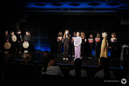 Trzy dziewczynki rozmawiają z przodu sceny. Za nimi krajobrazy pustynne. Po lewej stronie dzieci ubrane na czarno trzymają kolorowe zegary.