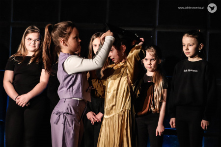 Dziewczynka w fioletowym kostiumie zmienia nakrycie głowy dziewczynce w złotej sukience. Z tyłu dzieci ubrane na czarno przyglądają się sytuacji.