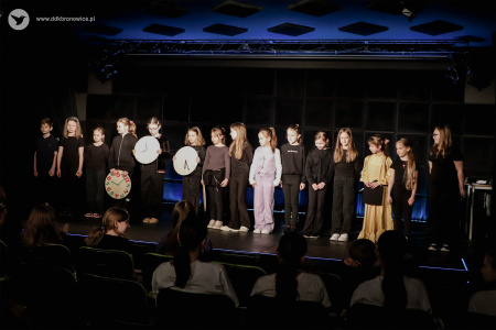 Cała grupa stoi na scenie. Po lewej stronie widać dzieci trzymające zegary.