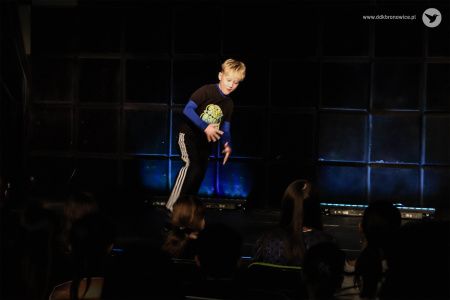 Chłopiec energicznie tańczy na środku sceny. Za nim niebieskie światło.