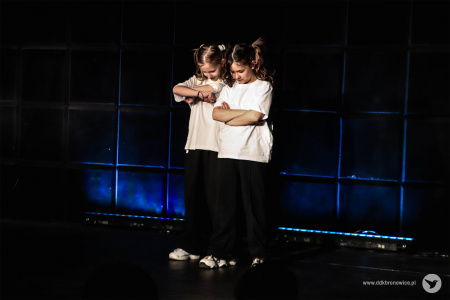 Na scenie stoją dwie dziewczynki. Głowy mają skierowane do dołu i maja założone ręce. Ubrane są w białe koszulki i czarne spodnie.