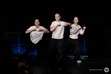 Trzy starsze dziewczyny tańczą w białych koszulkach i czarnych spodniach. Ręce mają ułożone na wysokości ramion.