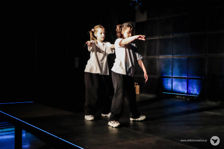 Dziewczynki stoją bokiem do widowni. Mają wyciągnięte ręce, energicznie tańczą.