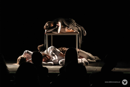 Aktorzy leżą pod małym stołem. Na stole leży aktorka. Ubrani są w białe kostiumy. Oświetla ich światło boczne.