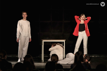 Na środku sceny stół. Pod nim aktorka i aktor. Po lewej stronie aktor w białym stroju. Po prawej stronie aktorka w czerwonej marynarce.