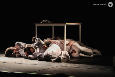 Aktorzy w białych strojach leża pod małym stołem na środku sceny.