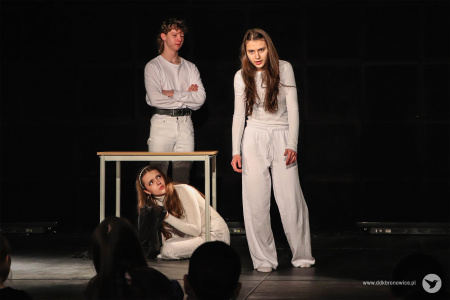 Na środku stół. Pod nim aktorka. Obok stołu stoi aktorka. Za stołem stoi aktor.