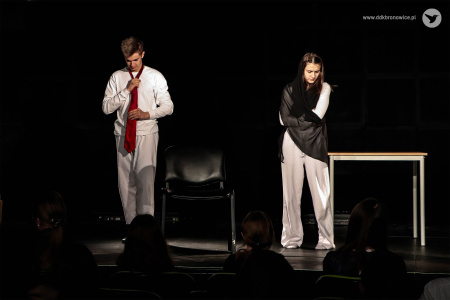 Aktor po lewej w białym stroju zakłada czerwony krawat. Aktorka po prawej stronie zakłada czarny szal.