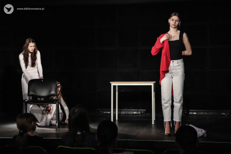 Dwie aktorki w białych kostiumach stoją po lewej stronie. Jedna z nich chowa się za krzesłem. Aktorka po prawej stronie zakłada czerwona marynarkę.