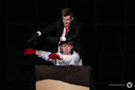 Aktor w białym stroju ma założone czerwone rękawiczki. Za nim stoi aktor w czarnej marynarce i czerwonym krawacie i czarnych rękawiczkach.