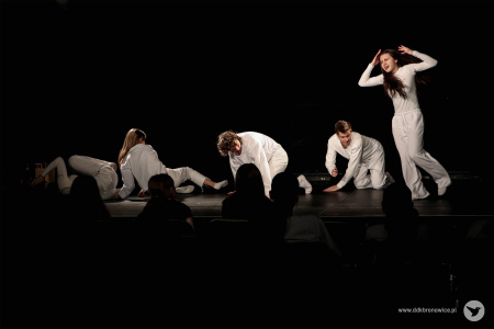 Czwórka aktorów w białych strojach leży na scenie. Po prawej stronie stoi aktorka w białym kostiumie.