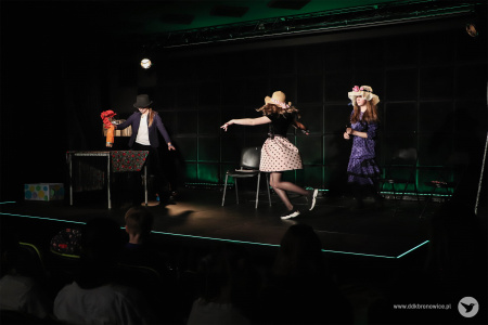 Dwie dziewczynki ubrane w sukienki w groszki skaczą po prawej stronie sceny. Po lewej stronie, obok stolika z obrusem stoi dziewczynka w czarnej marynarce i kapeluszu.