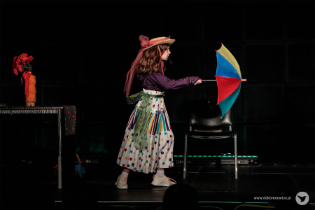 Dziewczynka w jasnej spódnicy w zielone groszki trzyma kolorową parasolkę. Na głowie ma słomiany kapelusz.