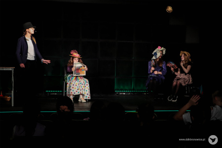 Dziewczynka w spódnicy w kolorowe groszki siedzi na fotelu. Po prawej stronie sceny widać dwie dziewczynki siedzące na krzesełkach.