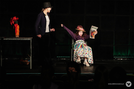 Dziewczynka w jasnej spódnicy w kolorowe groszki siedzi na fotelu i czyta gazetę. Obok niej stoi dziewczynka w czarnej marynarce i kapeluszu.