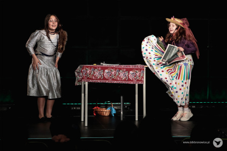 Dwie dziewczynki tańczą przy stoliku. Dziewczynka po lewej stronie na założoną srebrną sukienkę. Po prawej stronie stoi dziewczynka w jasnej sukience w kolorowe groszki.