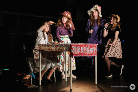 Cztery dziewczynki ubrane w kolorowe spódnice w groszki stoją wokół stołu. Jedna z nich siedzi na krześle przy stole.