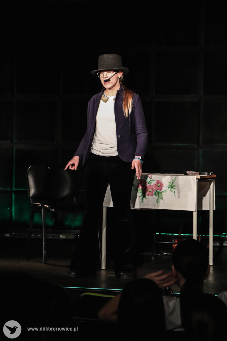 Dziewczynka w czarnej marynarce i kapeluszu mówi swoja kwestię. Stoi na środku sceny przy stoliku z białym obrusem.