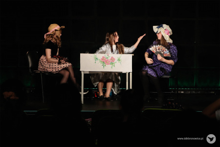 Przy stoliku siedzą trzy dziewczynki. Na środku siedzi dziewczynka w srebrnej sukience. Na stoliku biały obrus.