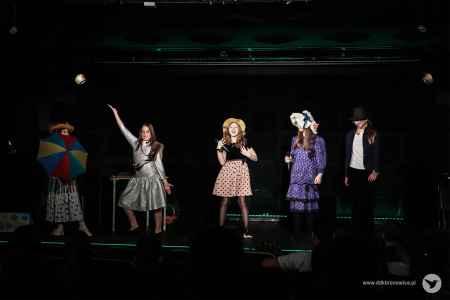 Dziewczynki w kolorowych spódnicach tańczą z przodu sceny. Na środku stoi dziewczynka w jasnej spódnicy w kolorowe groszki. Ma na głowie słomiany kapelusz z kolorową kokardką.