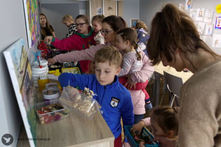 Uczestnicy warsztatów w salce. Kobieta w beżowym swetrze podaje materiały warsztatowe małej dziewczynce. Obok nich stoi chłopak w niebieskiej bluzie. W tle widać kobiety, które pobierają potrzebne materiały do stworzenia swojej ozdoby.