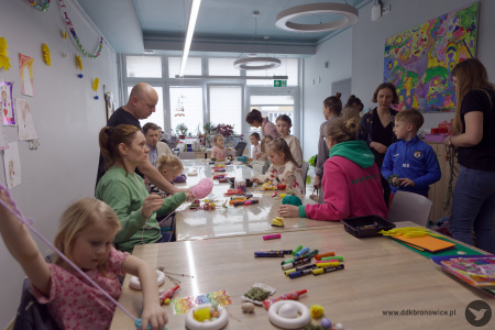 Uczestnicy tworzą swoje świąteczne ozdoby wielkanocne przy długim stole. Na stole rozłożone flamastry i artykuły papiernicze oraz materiały artystyczne.