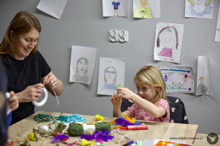 Przy stole warsztatowym siedzi dziewczynka. Ubrana jest w różową koszulkę. Wykorzystuje kolorowe nici i materiały do swojej ozdoby świątecznej.