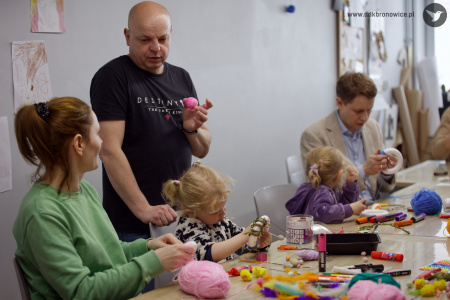 Mężczyzna w czarnej koszulce pokazuje dorosłej kobiecie różową pisankę. Obok mężczyzny siedzi mała dziewczynka.