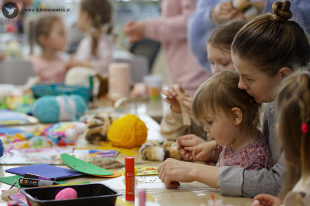 Mama trzyma na nogach małą córeczkę. Siedzą przy stole warsztatowym i wspólnie tworzą ozdobę świąteczną.