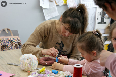 Kobieta w beżowym swetrze pomaga córce w skonstruowaniu kolorowej ozdoby świątecznej. Na stole widać wielką białą włóczkę.