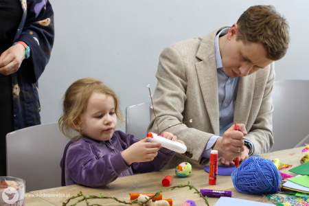 Mężczyzna w jasnej marynarce tworzy fragment artystycznej ozdoby. Obok niego dziewczynka tworzy inny element ozdoby.