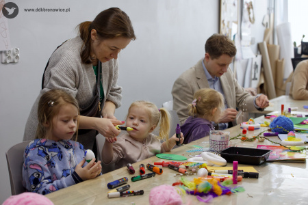 Kobieta w szarym swetrze pomaga dziewczynce w stworzeniu ozdoby. Kobieta stoi nad siedzącą dziewczynką. Przed nimi na stole leżą włóczki, kleje i materiały.