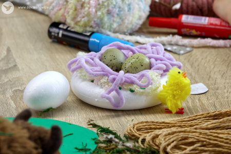 Na okrągłym elemencie leżą fioletowe wstążki. Na wstążkach widać jajka. Obok ozdoby świątecznej stoi żółty kurczaczek.