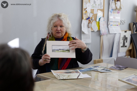 Prowadząca warsztaty pokazuje uczestnikom obraz w białej ramce. Prowadząca ubrana jest w czarną bluzę i pomarańczowy szal.