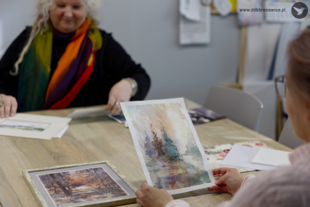 Uczestniczka ogląda obraz namalowany akwarelą. W tle, za stołem siedzi prowadząca warsztaty.