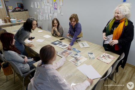 Pięć uczestniczek warsztatów siedzi przy stole. Trzy siedzą po lewej stronie. Dwie zajęły miejsca pop przeciwnej stronie. Obok stołu stoi prowadząca.