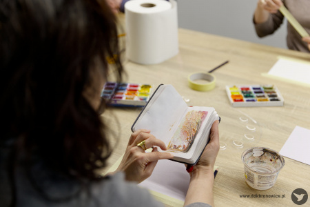 Uczestniczka ogląda notes. Na małych kartkach widać małe obrazy akwarelowe. Na stole, w tle widać farby i pędzle.