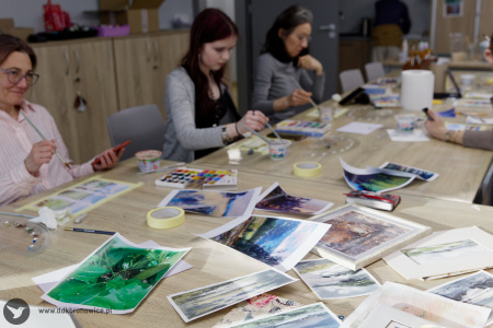 Na stole rozłożone obrazy. Wszystkie są malowane technika akwareli. Papier nabrał charakterystycznych zagięć od wody. W tle trzy uczestniczki malują swoje obrazy.