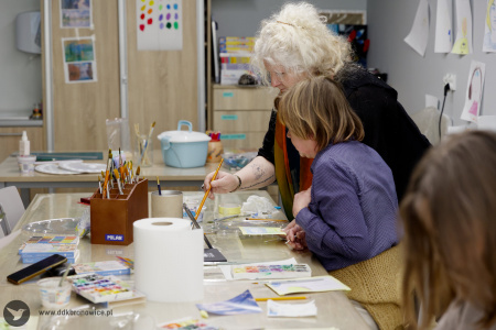 Prowadząca warsztaty z akwareli pomaga uczestniczce w malowaniu. Uczestniczka ma niebieską koszulę. Prowadząca trzyma pędzel. Uczestniczka przygląda się.
