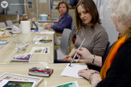 Prowadząca pomaga uczestniczce w malowaniu. Uczestniczka ma szary golf i przygląda się prowadzącej, która maluje na białej kartce.