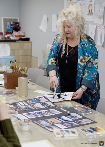 Prowadząca układa obrazy na stole. Ma założoną kolorową tunikę.